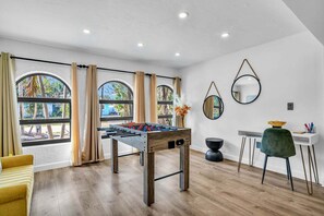 Una habitación con mesa de fútbol, escritorio y silla, y ventanas amplias con cortinas.