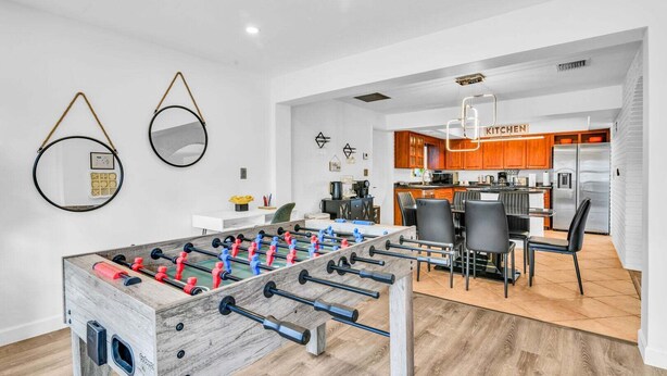 A modern kitchen with wooden cabinets, a stainless steel refrigerator, and a foosball table in a bright room with white walls and wooden flooring. - 1750 Managed by Brampton Park (Pompano Beach)