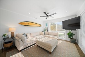 Un salon comprenant un canapé d’angle, un ventilateur de plafond, une planche de surf accrochée au mur, un téléviseur et une grande fenêtre.