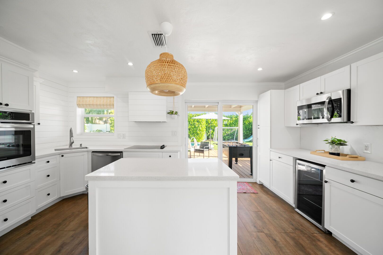 Une cuisine moderne avec des meubles blancs, un îlot central, un luminaire suspendu et une vue sur la terrasse extérieure.