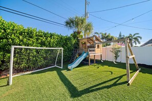 Une cour arrière clôturée avec une pelouse artificielle, un but de football, une cabane en bois avec un toboggan et un ensemble de balançoires.