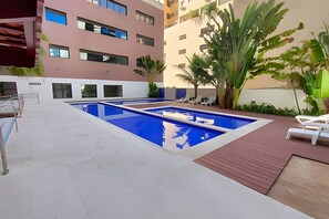 A rooftop pool area with blue water, white lounge chairs, and palm trees.