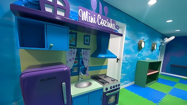 Una cocina de juguete para niños con un refrigerador morado, una estufa y un fregadero. La habitación tiene una decoración azul y verde con un tapete de juego en el suelo.