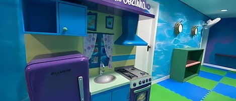Una cocina de juguete para niños con un refrigerador morado, una estufa y un fregadero. La habitación tiene una decoración azul y verde con un tapete de juego en el suelo.