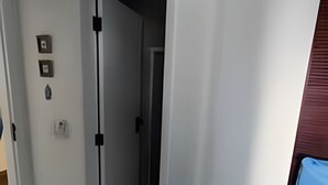 Un pasillo con una puerta que da a otra habitación. Hay tres interruptores de luz en la pared y un cubo de basura en la esquina.