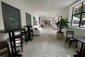 A spacious corridor with a long table and chairs on the left, a white sofa and a potted plant on the right, and a fire extinguisher on the wall. - SHB Lazer completo no melhor do Guarujá (Guarujá)