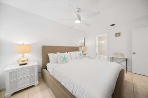 A bedroom with a bed, a nightstand, a lamp, a ceiling fan, a bathroom door, and a small table with a sign. - 2219 Managed by Brampton Park (Delray Beach)