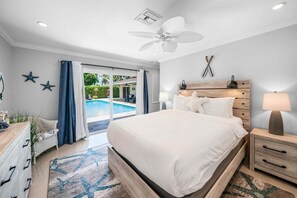 Une chambre avec un grand lit, un ventilateur de plafond, une table de chevet avec une lampe et une vue sur une piscine, visible à travers une porte vitrée.