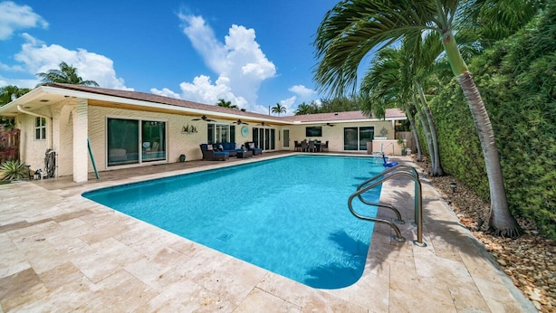 Ein Schwimmbad mit einer Terrassenfläche, einer überdachten Sitzgruppe und einem Haus mit großen Fenstern und einem Balkon.