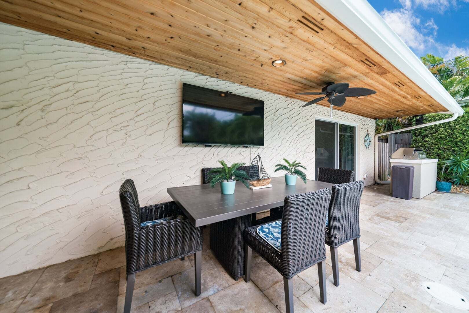 An outdoor patio with a table and chairs, a ceiling fan, and a flat-screen TV mounted on the wall.