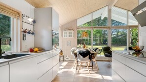 Une cuisine moderne avec des meubles blancs, un coin repas doté d’une chaise noire et de grandes fenêtres offrant une vue sur l’extérieur.