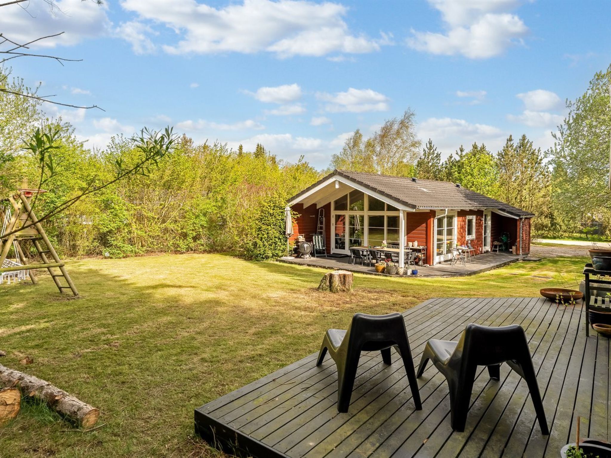 Une terrasse en bois avec des chaises, un petit bâtiment doté d’un porche et une échelle dans une zone herbeuse bordée d’arbres.