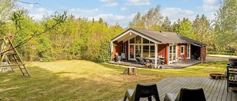 A wooden deck with chairs, a small building with a porch, and a ladder in a grassy area with trees.