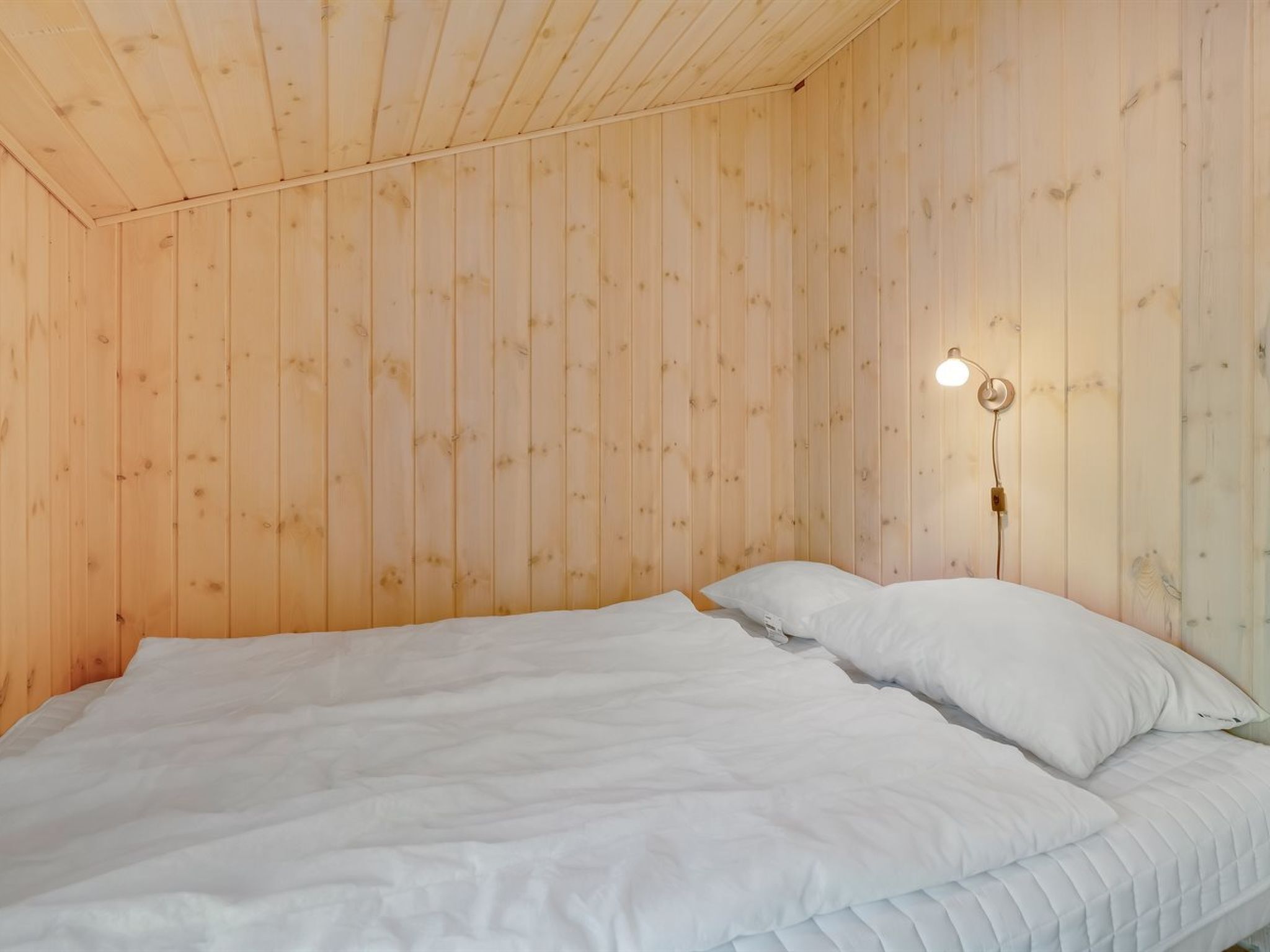 Una habitación con paredes de madera, una cama con ropa blanca y una lámpara de pared.