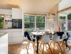 Eine moderne Küche mit weißen Schränken, ein Essbereich mit Tisch und Stühlen sowie große Fenster mit Blick ins Freie.