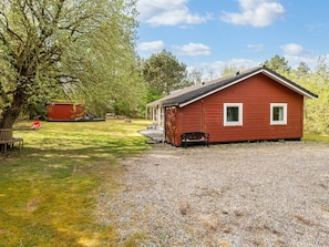 A red wooden cabin with a gravel driveway, a bench, and a red shed in a grassy area with trees.