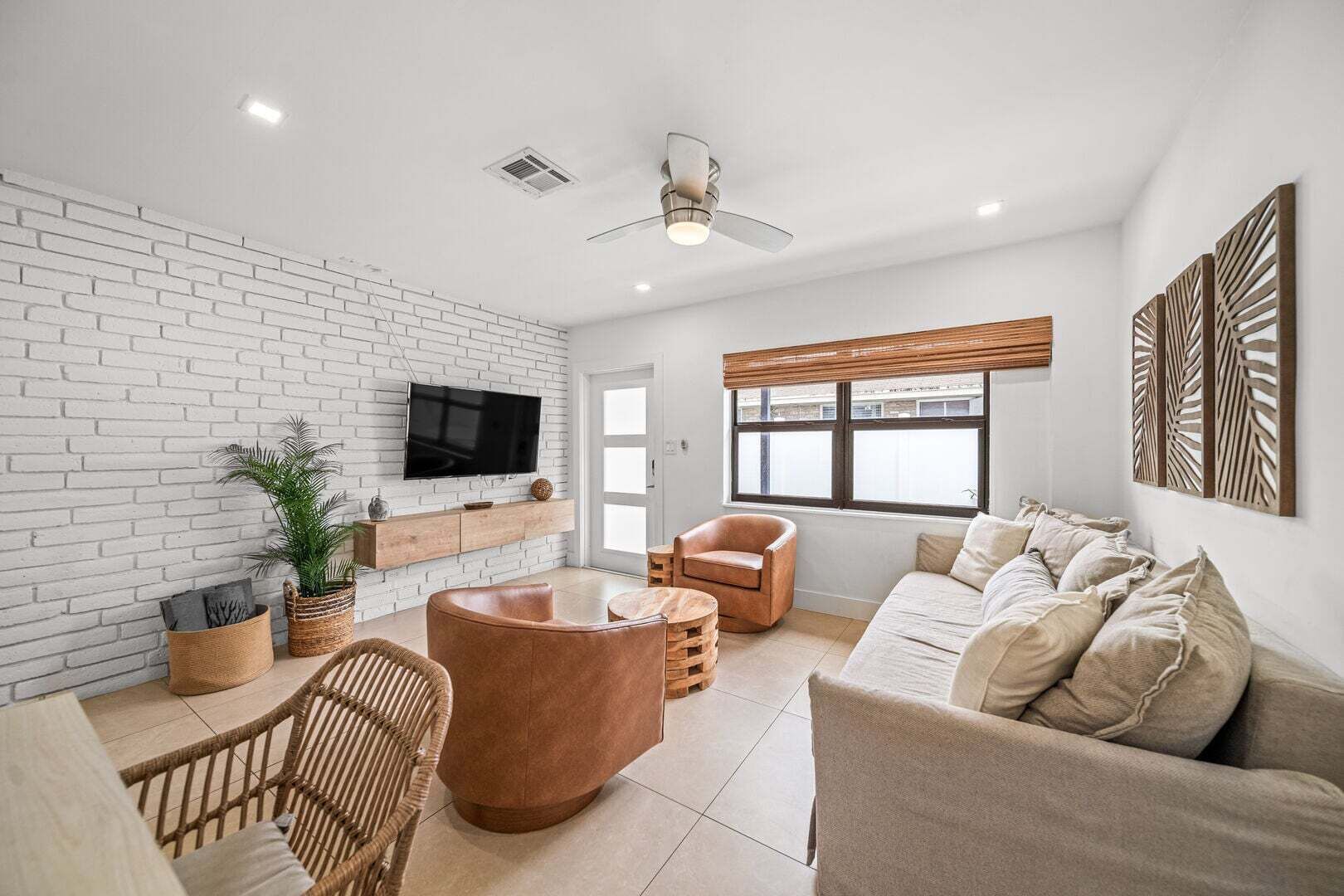 A modern living room with a white brick wall, a flat-screen TV, a large window with blinds, a brown leather sofa, a wooden coffee table, and a wicker chair.