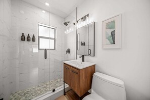 Une salle de bain moderne avec une douche à l’italienne en verre, un meuble-lavabo en bois, des toilettes blanches et un tableau abstrait encadré accroché au mur.