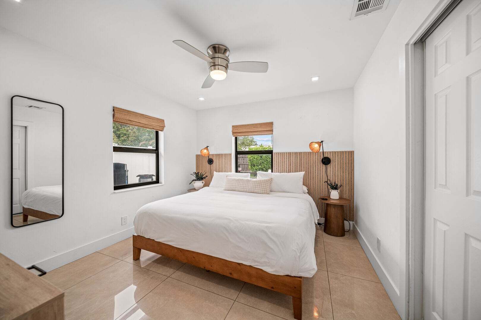A bedroom with a bed, a ceiling fan, two bedside tables with lamps, a large mirror, and a window with blinds.