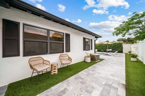 Une maison moderne avec une façade blanche, des volets noirs et une terrasse pavée agrémentée de deux fauteuils en osier et d’une petite table ronde.