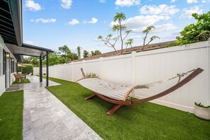 A hammock in a backyard with a white fence, a paved walkway, and a house with a covered patio.