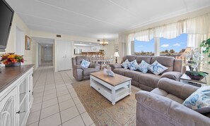 Ein Wohnzimmer mit einer Sofagarnitur, einem Couchtisch, einem Fernseher und einer Küche im Hintergrund.