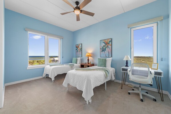 Ein Schlafzimmer mit zwei Betten, einem Schreibtisch, einem Stuhl, einem Deckenventilator und großen Fenstern mit Blick aufs Meer.