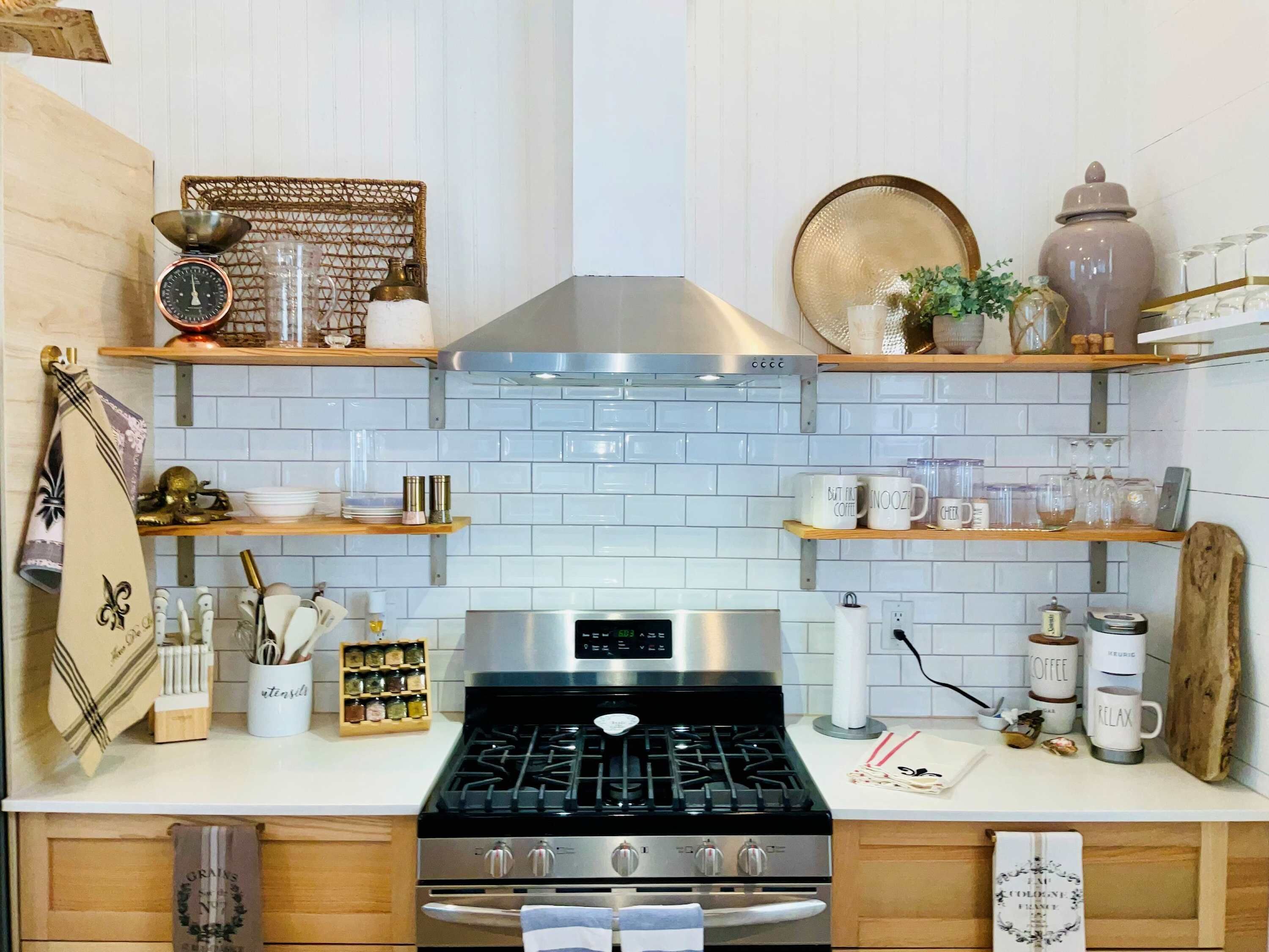 Una cocina con salpicadero de azulejos blancos, electrodomésticos de acero inoxidable, estanterías de madera y diverso menaje de cocina.