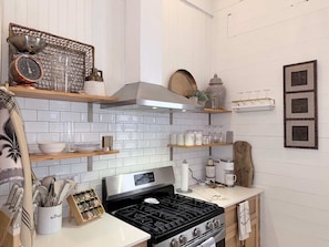 Cocina con salpicadero de azulejos blancos, electrodomésticos de acero inoxidable, repisas de madera y objetos decorativos.