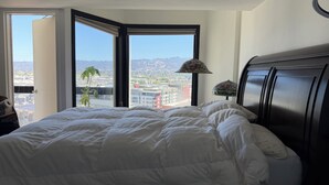 A hotel room with a large bed, a nightstand with a lamp, and a view of a cityscape through a corner window. - Cozy 1-bedroom condo 4miles from uc Berkeley with lots of public transportation (Emeryville)