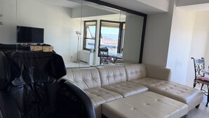 A modern living room with a large sofa, a television, and a dining area visible through a large window. - Cozy 1-bedroom condo 4miles from uc Berkeley with lots of public transportation (Emeryville)