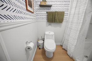 Pieni kylpyhuone, jossa on wc, rulla wc-paperia, roskakori ja pyyhe, joka on ripustettu seinälle.