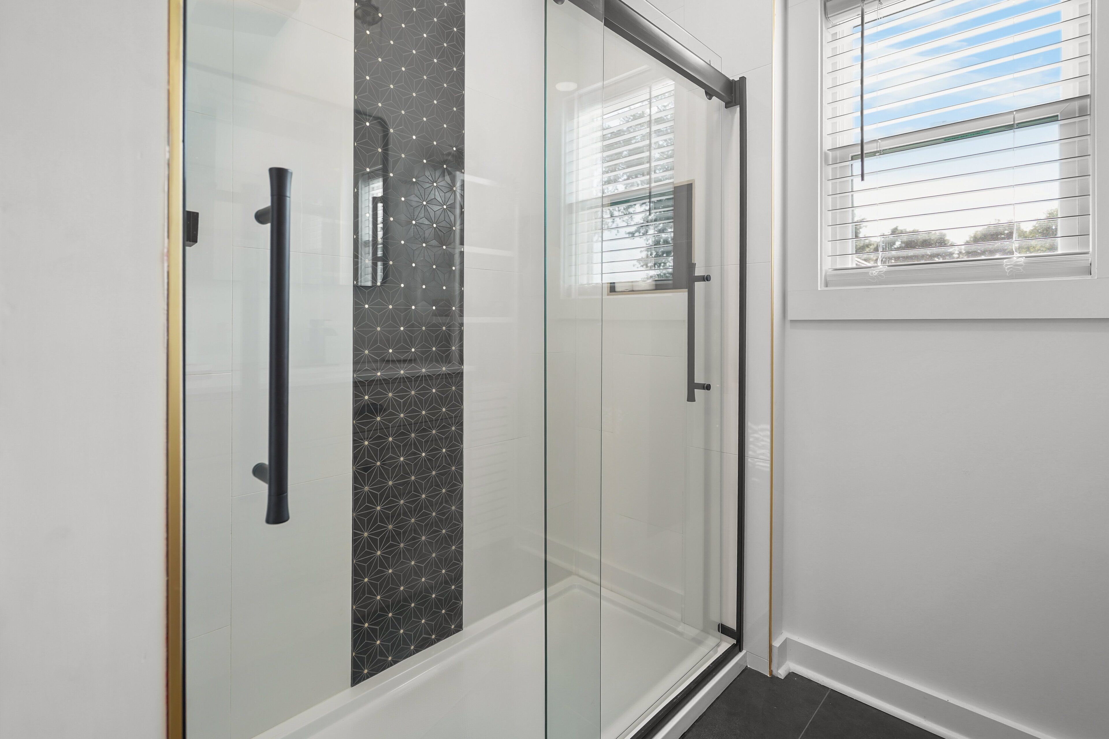 A modern bathroom with a glass shower enclosure, a white tiled wall, and a window with blinds.