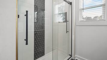 A modern bathroom with a glass shower enclosure, a white tiled wall, and a window with blinds.