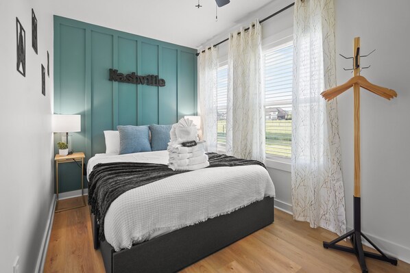A bedroom with a teal accent wall, a bed with a white and gray blanket, a bedside table with a lamp, and a view of the outdoors through a window with sheer curtains.