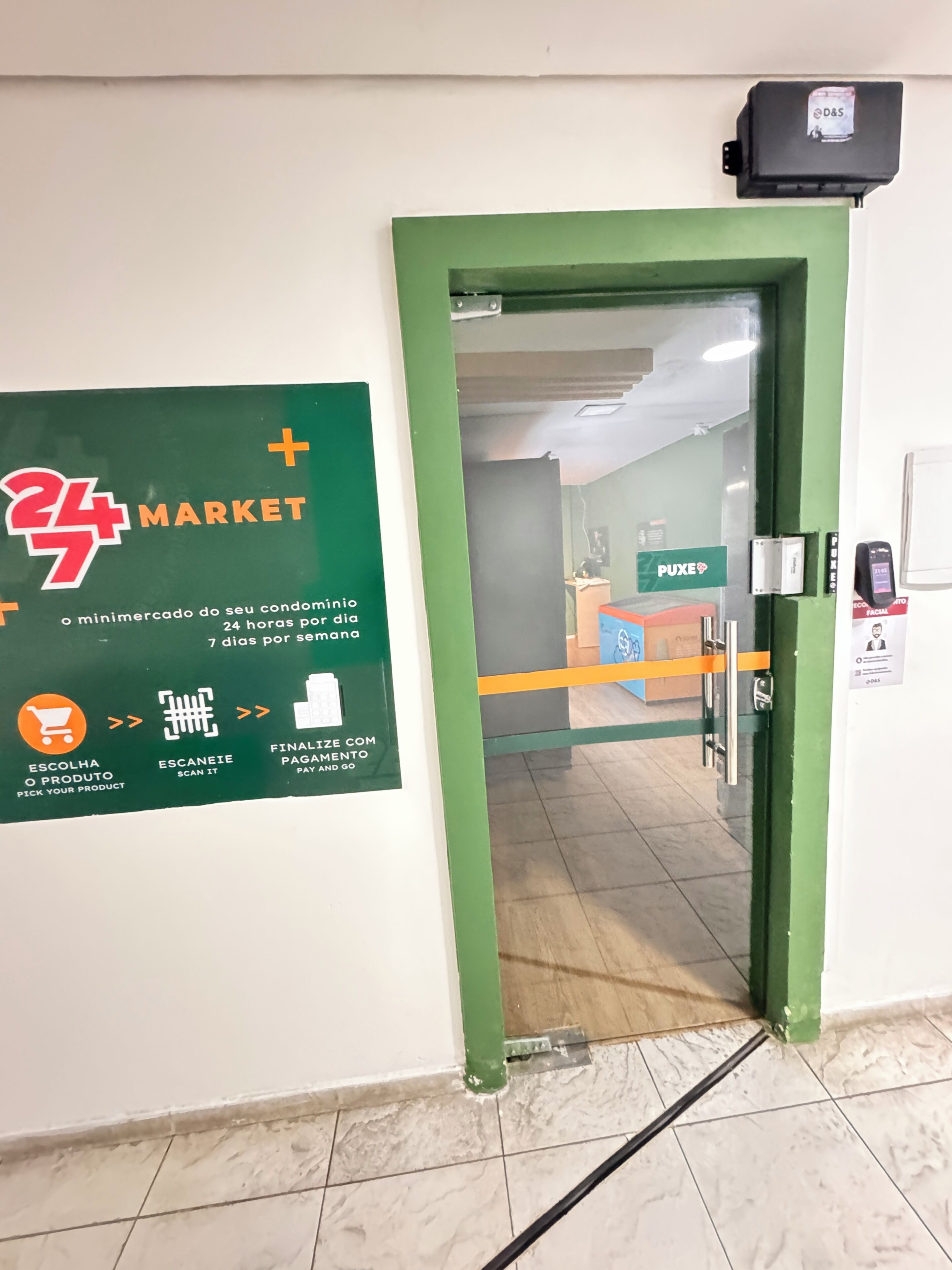 Porta verde com moldura que dá para uma área de mercado, com um aviso indicando serviço 24 horas por dia.
