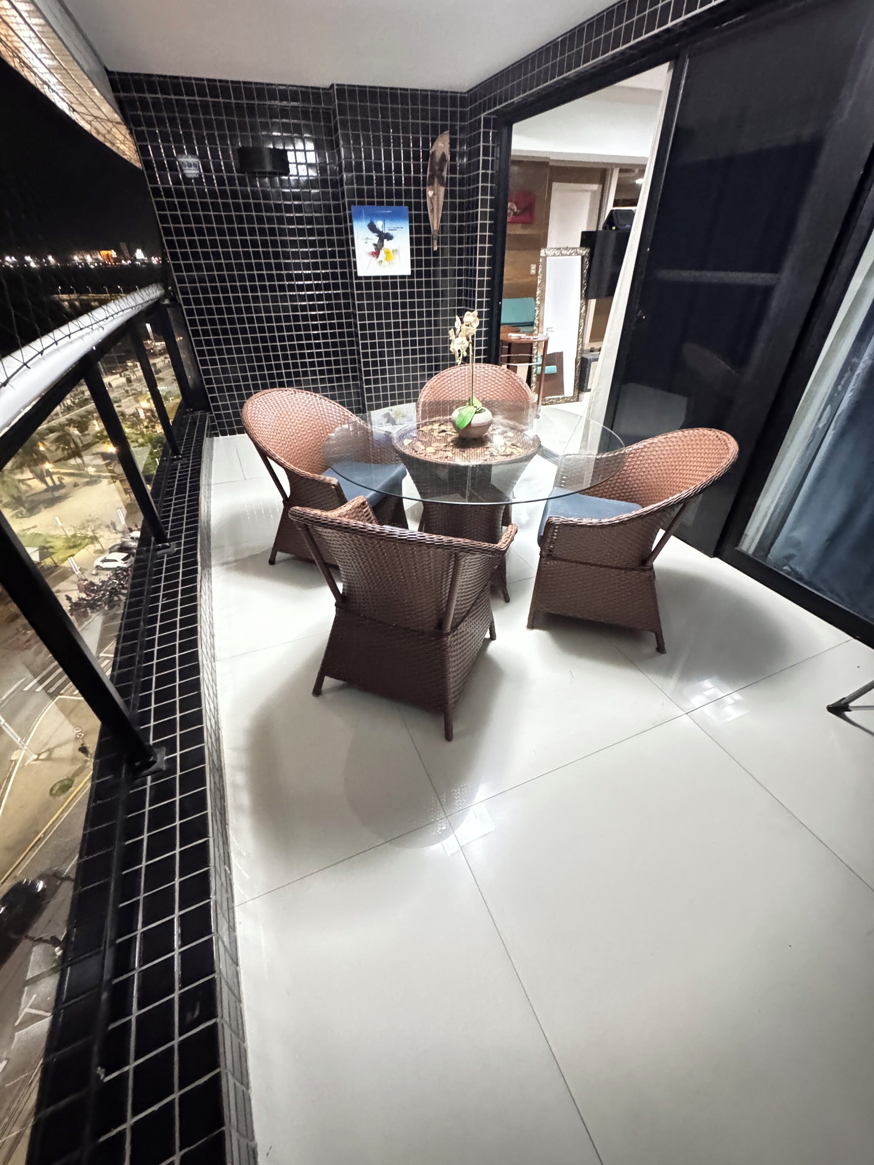 A balcony with a glass table and wicker chairs, a tiled wall, and a view of the street below.