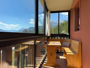 Ein Balkon mit einer hölzernen Bank und einem Tisch, großen Fenstern mit Blick auf Berge und einer roten Wand.