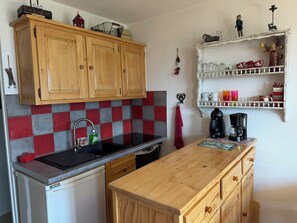 A kitchen with wooden cabinets, a red and grey tiled backsplash, a sink, a coffee maker, and a shelf with decorative items. - Balcon Panoramique • Vue Montagne • Barcelonnette (Barcelonnette)