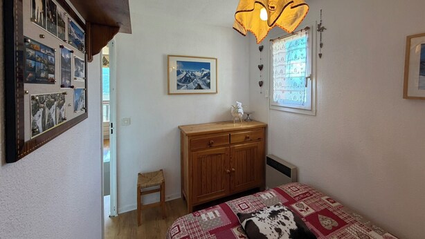 Ein Schlafzimmer mit einem Bett, einem Holzschrank, einem kleinen Stuhl, einem gerahmten Bild an der Wand und einem Fenster mit Vorhang.
