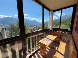 Ein Balkon mit Holztisch und Bank, der Blick auf Berge und eine Stadt gewährt.