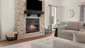 A living room with a stone fireplace, a flat-screen TV mounted above it, a sofa, and a clock on the wall. - Not So Crappie Cabin - 3BR/3BA, Private Hot Tub, Resort Pools & Lazy River (Hollister)