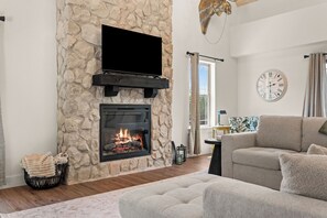 A living room with a stone fireplace, a flat-screen TV mounted above it, a sofa, and a clock on the wall. - Not So Crappie Cabin - 3BR/3BA, Private Hot Tub, Resort Pools & Lazy River (Hollister)