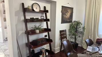 Una sala de estar con una estantería de madera, un cuadro, una planta en maceta y un comedor con sillas y una mesa.