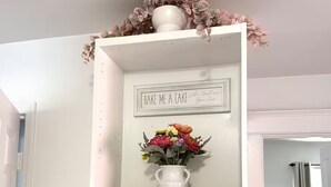 Ein weißes Regal mit einem Schild, einer Vase mit Blumen und farbenfrohen Gläsern.