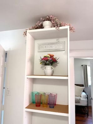A white shelf with a sign, a vase with flowers, and colorful glasses.