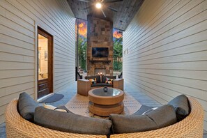 A covered outdoor seating area with a stone fireplace, wicker furniture, and a flat-screen TV mounted on the wall. - The Gather House | Private Pool • Sauna • Game Room • UTV Trail Access • Sleeps 26 (Broken Bow)