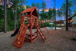 A wooden play structure with a slide and climbing wall, surrounded by trees and a building in the background. - The Gather House | Private Pool • Sauna • Game Room • UTV Trail Access • Sleeps 26 (Broken Bow)