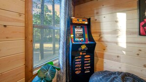 En Pac-Man-spelautomat i en stuga med träväggar och ett fönster med utsikt över träd.