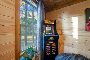 A Pac-Man arcade machine in a cabin with wooden walls and a window overlooking trees. - The Gather House | Private Pool • Sauna • Game Room • UTV Trail Access • Sleeps 26 (Broken Bow)